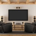 wall mounted tv with a speaker mounted above, 4 speakers placed on an entertainment unit underneath the tv. two black armchairs in front
